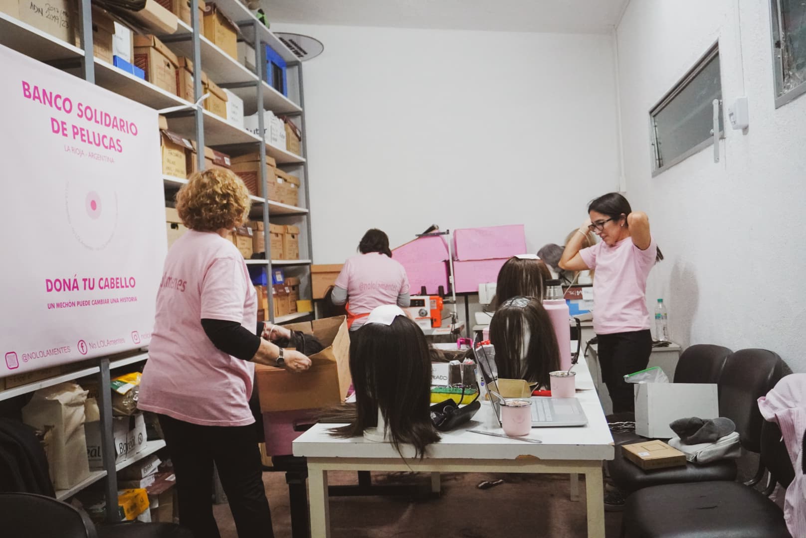 Voluntarias de noLOLAmentes con remeras rosas alrededor de la mesa de trabajo del taller, con cabezas de peluca y el banner del banco solidario de pelucas